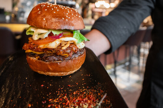 A close-up shot of a large gourmet burger with a sesame seed bun, lettuce, tomato, cheese, caramelized onions, and chili flakes on a black plate being served in a restaurant setting.