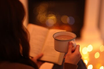 Woman with cocoa drink reading book near fireplace at home, closeup. Bokeh effect