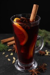 Tasty mulled wine in glass mug, spices and fir branches on black table, closeup