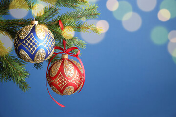 Obraz premium Christmas balls with pattern hanging on fir tree branches against blue background with blurred lights, closeup with space for text. Bokeh effect