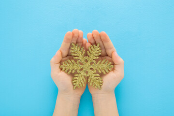 Little child opened palms holding and showing beautiful shiny golden snowflake covered with glitters on pastel blue table background. Closeup. Christmas decoration. Point of view shot. Top down view.