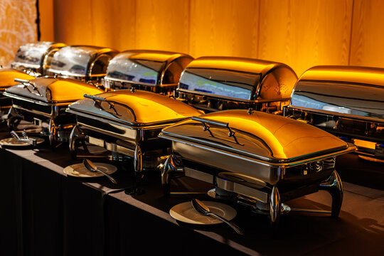 A row of shiny stainless steel chafing dishes on a black tablecloth, illuminated by warm orange lighting. Plates and serving spoons are neatly placed in front