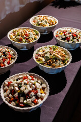 Six white scalloped bowls filled with assorted salads including Greek and Caesar styles, arranged on a purple tablecloth under warm lighting. Perfect for catering or buffet themes