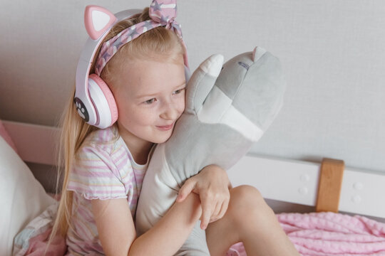 Young girl wearing pink cat-ear headphones and hugging gray stuffed cat on cozy bed. Happy child enjoying music in playful pink bedroom.