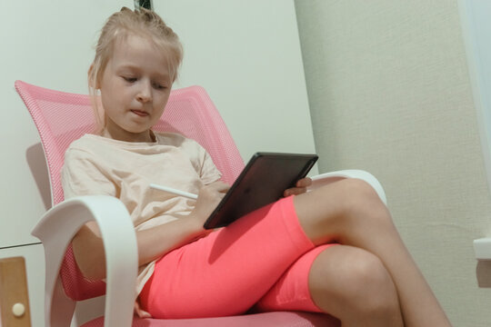 Young blonde girl focused on tablet in pink chair. Child engaged in digital learning or creative play at home with stylus. Indoor technology activity for kids - Powered by Adobe
