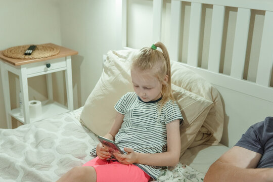 Young blonde girl and father using smartphones on cozy bed. Family bonding with technology in modern bedroom setting