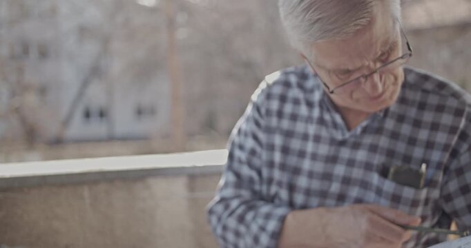 An older man with gray hair and glasses is engrossed in his reading or writing outdoors. The scene conveys a sense of peaceful focus and quiet contemplation for seniors.