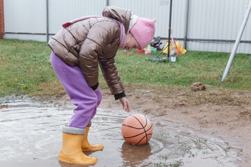 Young girl playing with a basketball in a muddy puddle after rain. Autumn outdoor activity for child. Fun and childhood concept.