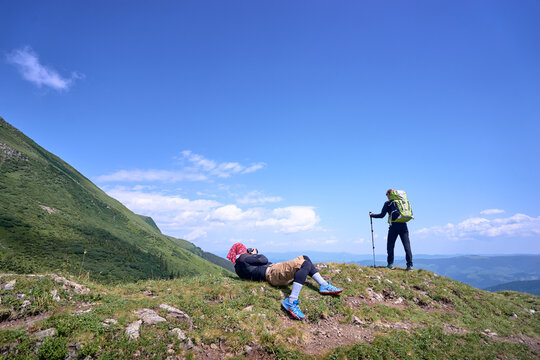 Hikers enjoy the scenic view from a mountain summit under clear blue skies. One is standing with gear while the other lies on the grassy ground, taking photo of the first one in tranquility of nature. - Powered by Adobe