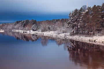 Serene winter landscape featuring a calm river reflecting snow-covered trees under a twilight sky, creating a tranquil atmosphere
