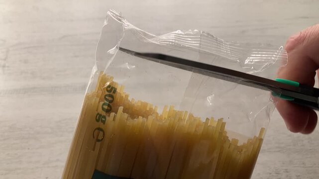 Getting Ready to Cook: A close-up of a person's hand using scissors to cut open a plastic package of dry, uncooked spaghetti in preparation for making an Italian meal