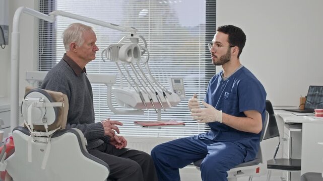 Young Middle Eastern male dentist speaking with Caucasian male senior patient seated in dental chair after completing treatment in bright modern clinic setting
