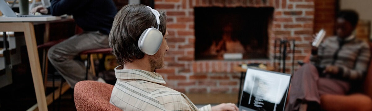 Website header shot of Caucasian young adult man wearing headphones working on laptop in modern business office, Black woman sitting in background using laptop near fireplace, open workspace setting