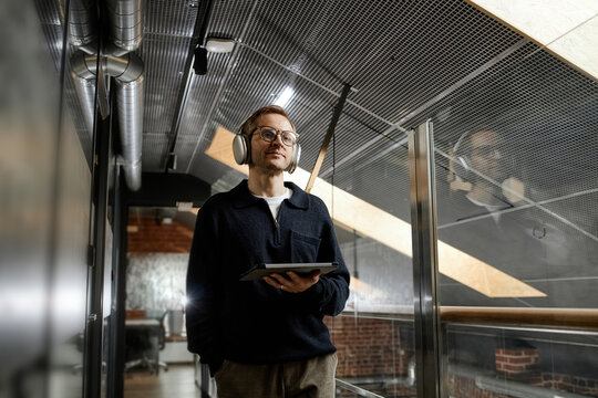 Caucasian young adult man wearing headphones holding digital tablet walking through modern business office corridor looking ahead with focused expression