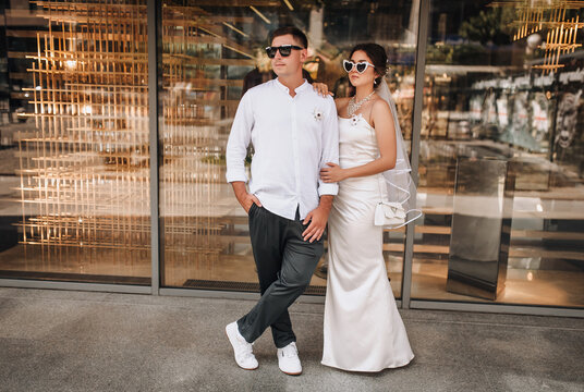 Wedding photography, portrait of happy cool newlyweds in sunglasses, young stylish groom and beautiful bride, standing against glass background of store on street in city. - Powered by Adobe