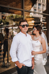 Wedding photography, portrait of happy cool newlyweds in sunglasses, young stylish groom and beautiful brunette bride, standing against glass background of store on street in city.