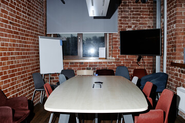 Empty modern business office meeting room featuring large conference table surrounded by multiple chairs, digital screen mounted on brick wall, flip chart standing near window