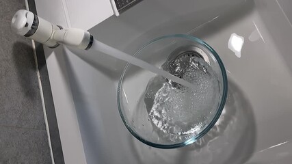 Pouring water from a tap into a glass bowlKitchen Preparations: A person fills a clear glass bowl with fresh, running water from a tap in a sink, a basic step for cooking or cleaning