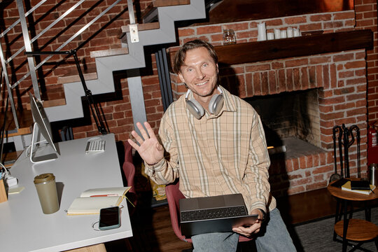 Portrait of middle aged Caucasian man smiling and waving while sitting in modern business office holding laptop with wireless headphones around neck, digital devices on desk nearby