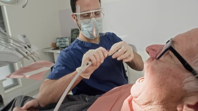 Dentist carefully treating senior patient teeth while using saliva ejector to maintain clean and clear working area in modern clinic environment