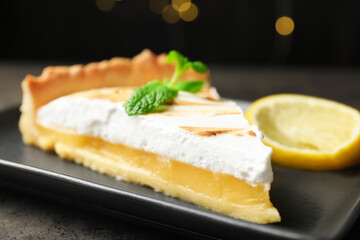 Slice of tasty lemon meringue tart with mint served on black table against blurred lights, closeup