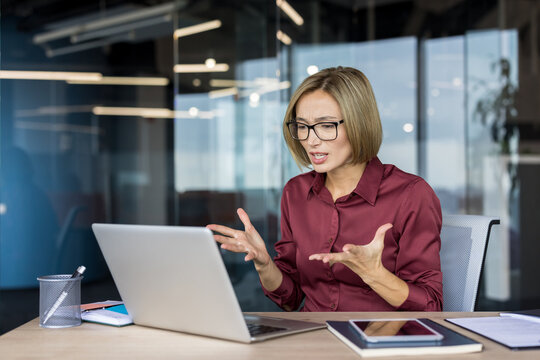 Businesswoman feeling frustrated and stressed, gesturing with her hands while having a challenging discussion or problem-solving during a video conference on her laptop in a modern office