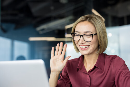 Businesswoman with glasses laughing and waving hand during a video conference on her laptop, communicating online while working in a modern office environment - Powered by Adobe