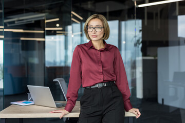 Young professional businesswoman standing confidently by her office desk, contemplating future business strategies within a modern corporate workspace setting