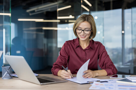 Businesswoman with glasses smiling while reviewing documents and notes, taking down details, and organizing information at her spacious, bright office workspace
