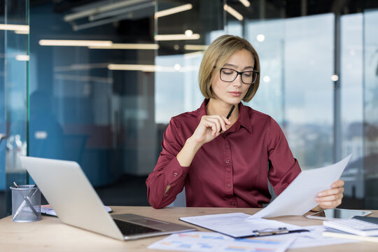 Businesswoman reviewing financial reports and data on paper, concentrating on her work at a desk with a laptop and various documents in a contemporary office environment