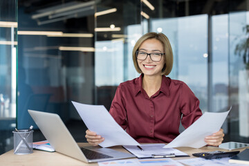Happy young businesswoman in glasses smiles at her desk, holding documents and working on a laptop in a modern office, confident and professional during her workday