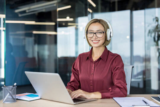 Smiling businesswoman wearing glasses and white headphones, sitting at an office desk and happily working on a laptop, embodying professionalism, concentration, and modern workplace communication
