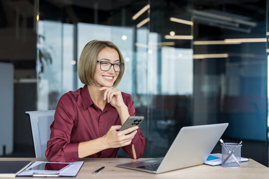 Smiling businesswoman at modern office desk holding smartphone and looking thoughtfully away, surrounded by laptop and tablet, conveying professional communication and success - Powered by Adobe