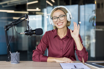 Smiling woman wearing headphones and glasses, sitting at a desk with a microphone, waving hand while broadcasting a live audio show online from a modern studio