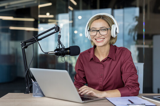 Young woman smiling while recording a podcast or live streaming content, wearing headphones and speaking into a professional microphone at a desk with a laptop and documents - Powered by Adobe