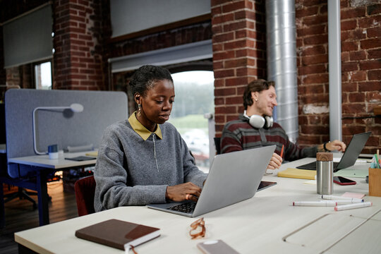 Black young adult woman working on laptop at modern business office desk, while Caucasian young adult man sitting in background using laptop with headphones around neck