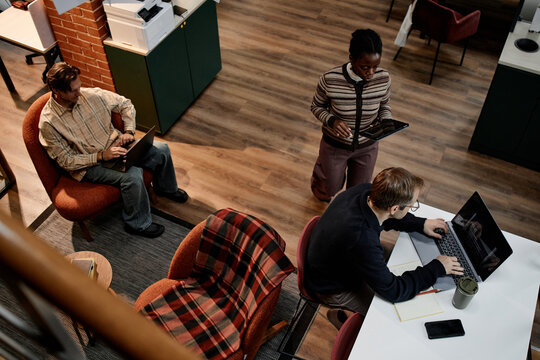 Middle aged Caucasian man sitting and typing on laptop, Black woman standing and holding digital tablet, middle aged Caucasian man working on laptop in modern business office