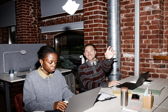 Young adult Black woman working on laptop, while young adult Caucasian man playfully throwing paper toward camera and smiling in modern business office with exposed brick walls