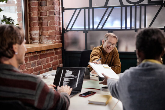 Young adult Caucasian man smiling while handing document to middle aged Black woman during business meeting, young adult Caucasian man using laptop with code on screen in modern office