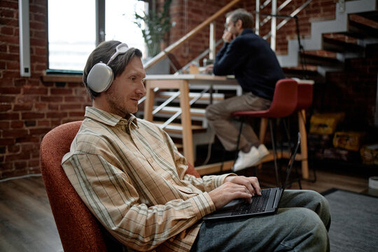 Caucasian young adult man wearing headphones working on laptop in modern business office, middle aged Caucasian man sitting at desk in background using desktop computer