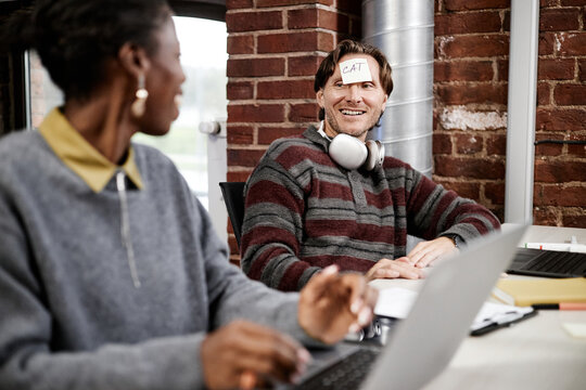 Caucasian young adult man sitting at desk with sticky note labeled cat on forehead smiling at Black woman colleague in modern business office, both using laptops during casual interaction