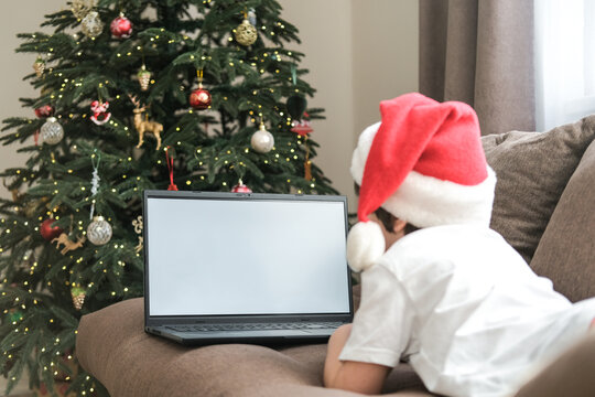 Boy in Santa hat using laptop with blank screen on sofa next to decorated Christmas tree. Concept: holidays, video calls, online shopping, remote learning, screen mockup