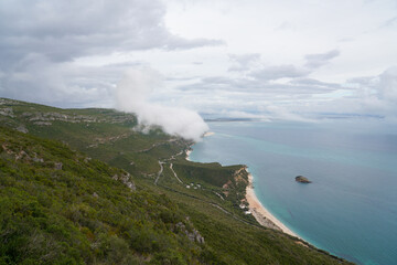 Winding coastal road in Portugal with misty green hills and turquoise sea. Rocky island near shore.