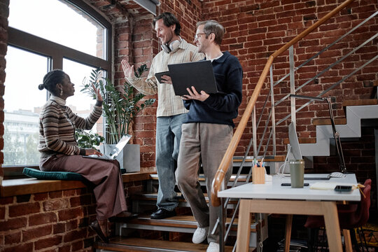 Multiethnic group of young adult and middle aged colleagues collaborating on laptops and digital tablet near window in modern business office, smiling and high fiving during discussion