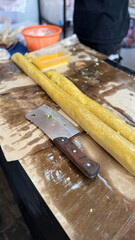 two long rolls of yellow ngohiong dough rest on a brown work surface beside a cleaver, with containers of seasoning and a vendor nearby, ready to be cut and cooked. he keng or ngo hiong rolls