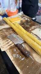 two long rolls of yellow ngohiong dough rest on a brown work surface beside a cleaver, with containers of seasoning and a vendor nearby, ready to be cut and cooked. he keng or ngo hiong rolls