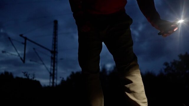 silhouette holding flashlight near tracks under dramatic sky and catenary poles, intense backlight and lens flare, lone figure in jeans, tense exploration vibe, cinematic night suspense