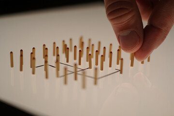 Hand Placing One of Many Wooden Dowels Arranged in Geometric Pattern on Illuminated White Surface, Creative Design