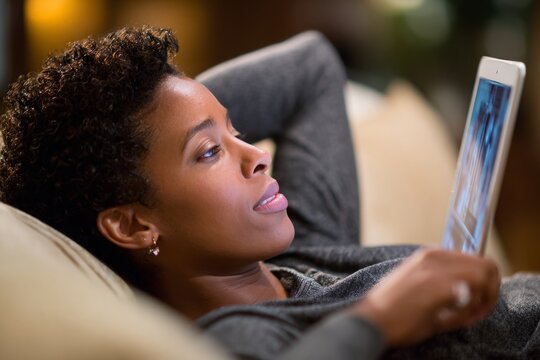 African American woman reclines on sofa, focused on digital tablet screen for entertainment. - Powered by Adobe