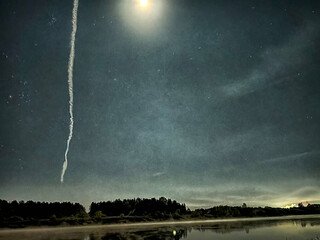Beautiful night sky above a tranquil lake with a bright moon and a contrail extending into the starry expanse